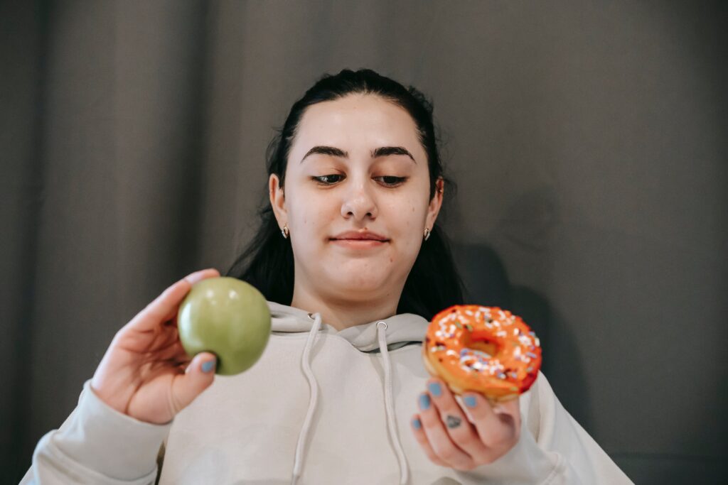 A picture of a girl with an apple and a donut. She seems to be trying to choose between a healthy apple and a delicious donut.