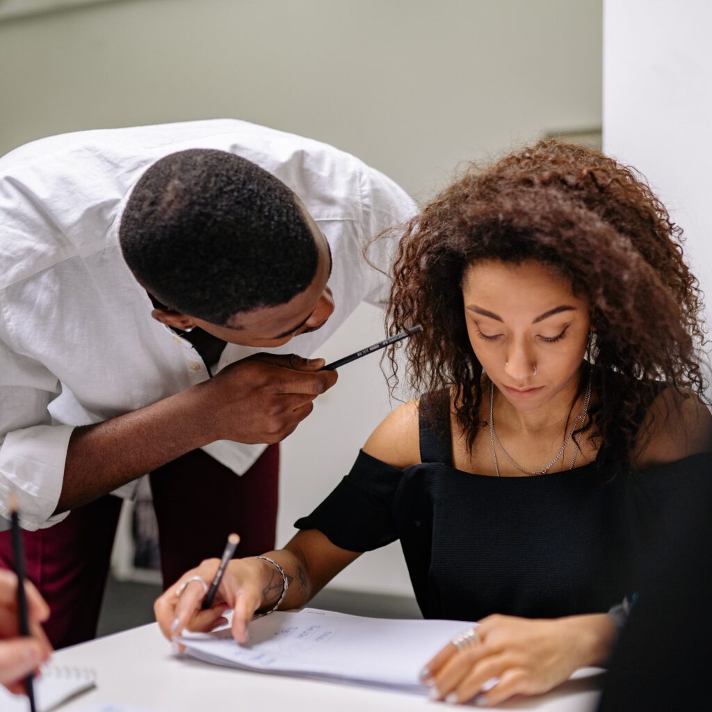 A woman siting down and a man standing over her, very close to her face and pointing at her. He seems angry and she seems afraid. Image is a symbol of workplace conflict.