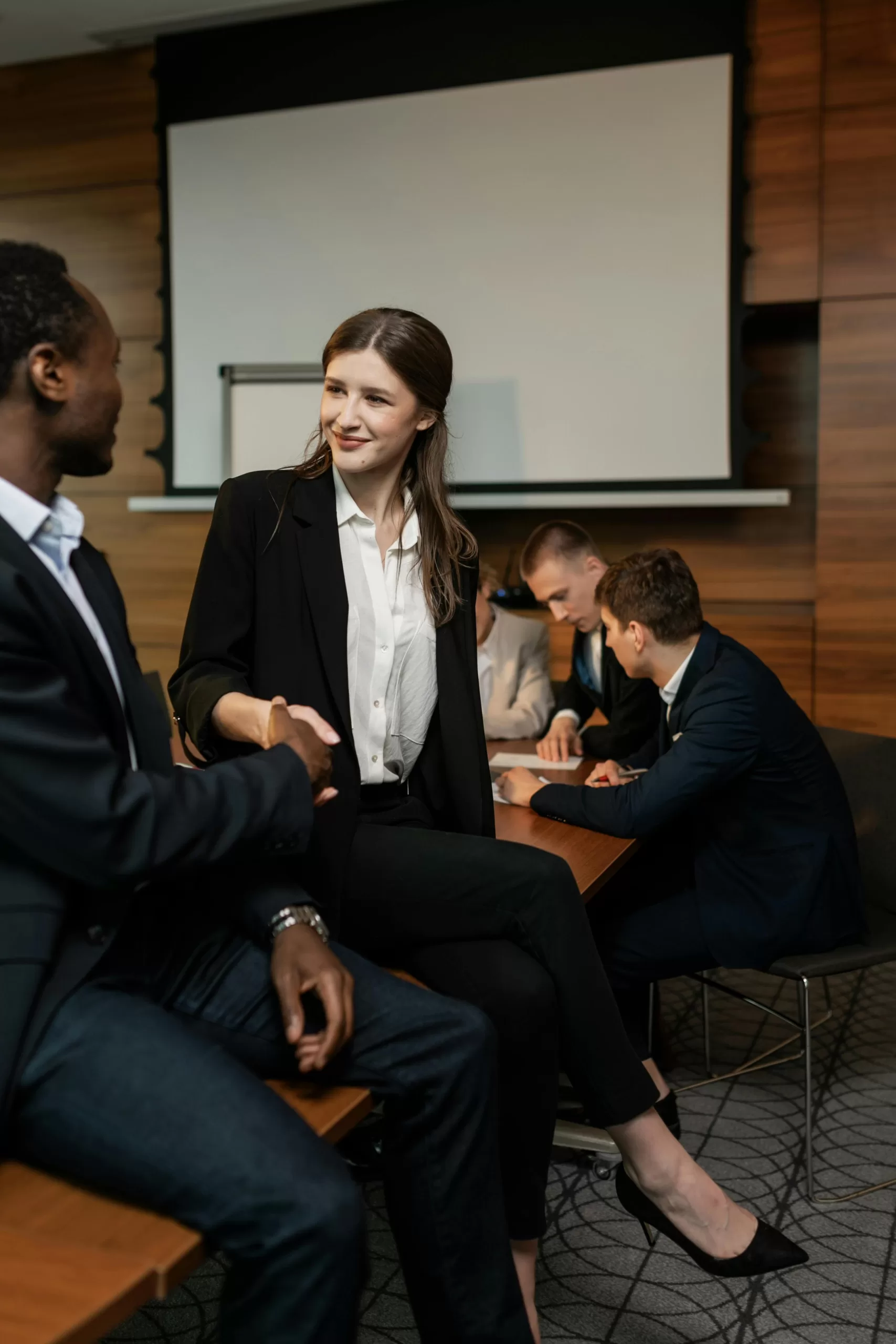 A well-dressed woman smiling and shaking hands with a man at a conference table, while three men collaborate on documents in the background, symbolizing teamwork and leadership.